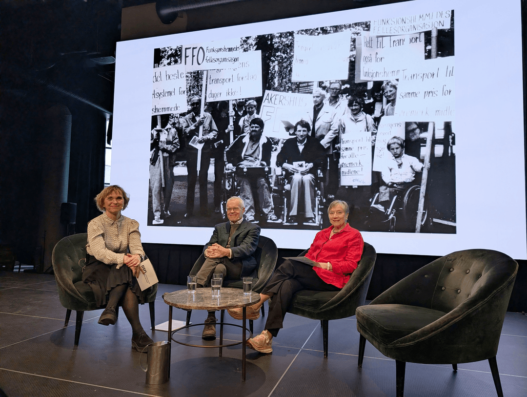 Tre personer sitter på en scene. Det er grønne fløyesstoler og et rundt bord med flere glass med vann. En kvinne til høyre har på seg rød skjorte og kort lyst hår. I midten sitter en mann i grønn dress han bruker briller. Til høyre sitter en dame med lyst kort hår. Hun har på seg lys skjorte og mørkt skjørt.