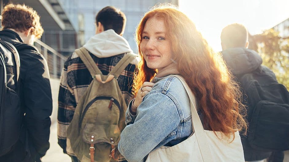 Fire studenter går ute og er på vei til forelesning. Foran går tre gutter med hver sin ryggsekk. Litt bak dem går en jente med en lys veske. Hun har rødt hår og blå denim jakke. Hun har ansiktet vent mot kamera og smiler.