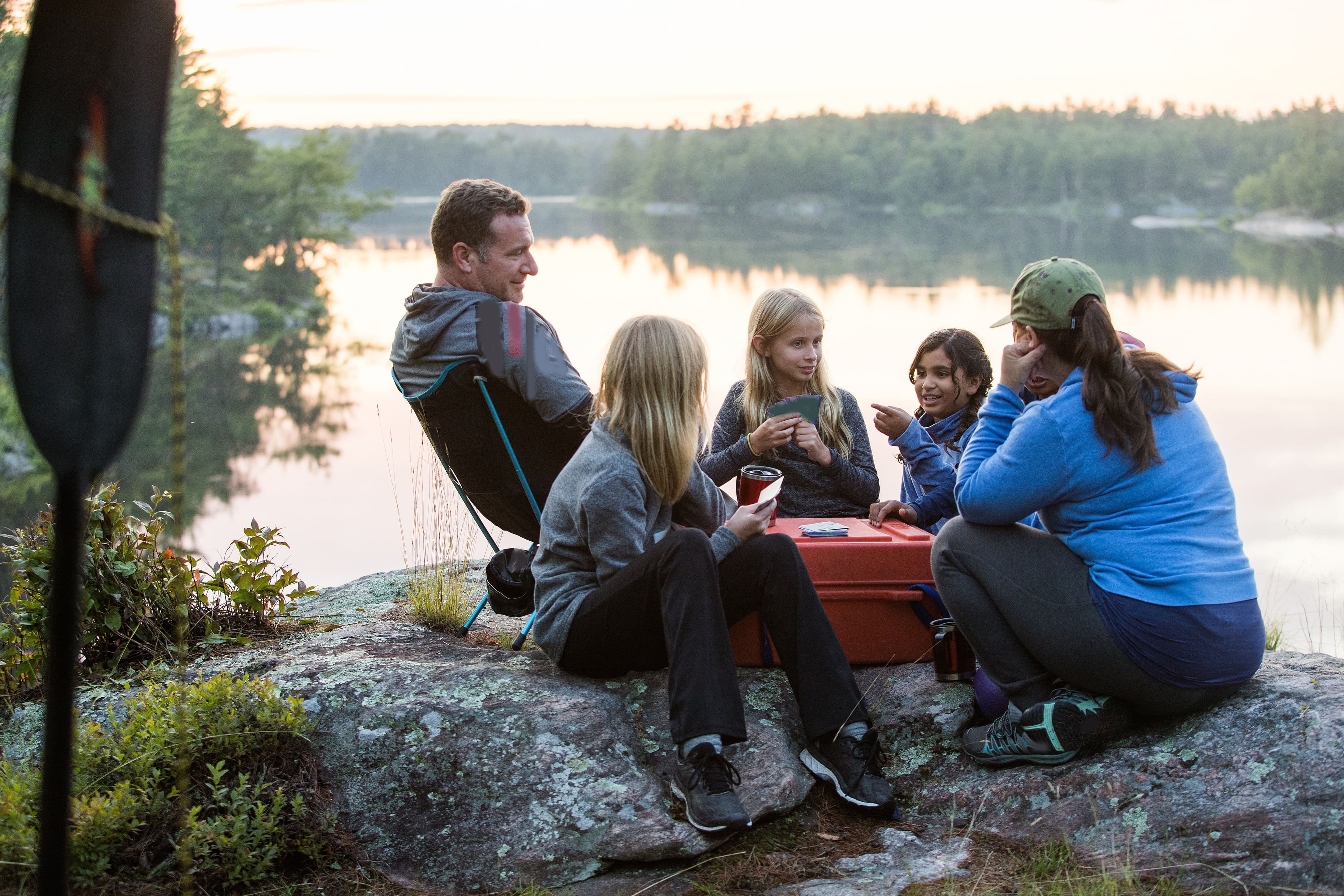 En familie på fem sitter ute på en sten, ut mot vannet. De snakker og ler, og sitter ved noe som ligner på en rød kasse.